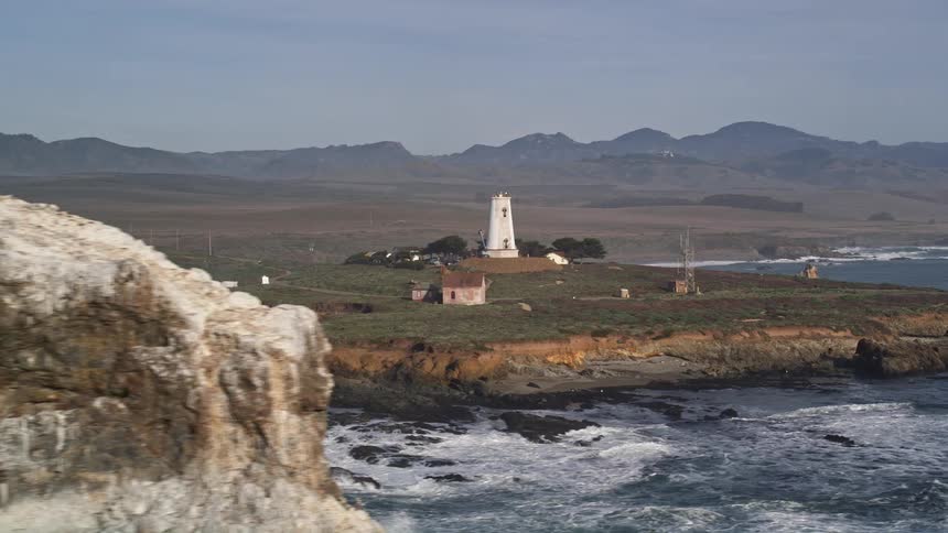 Flock of birds flies over ocean near shore with a lighthouse and other buildings