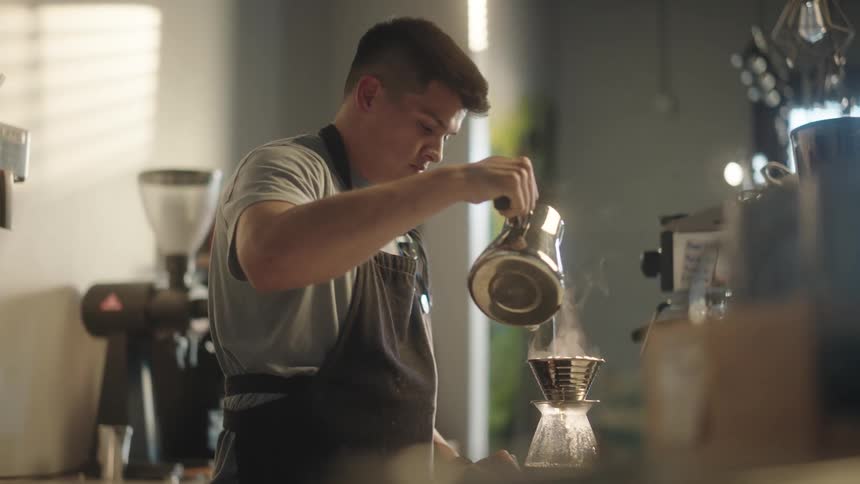 A young man is using a teapot to pour coffee into a glass container