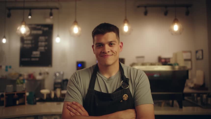 Portrait of a young man in an apron and arms crossed smiling while in a coffee shop