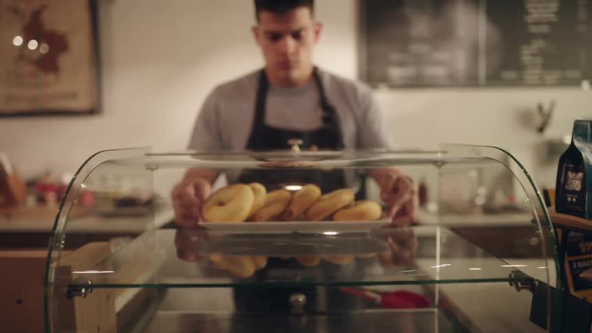 Man in an apron placing savory donuts inside a glass gland in a coffee shop