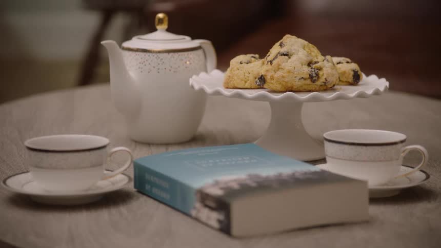 A teapot several cups and saucers a book and three scones on the table