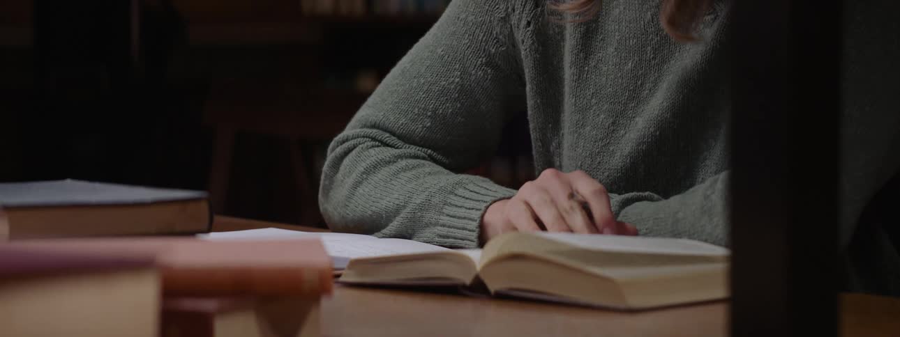 Hands of a woman sitting at the table reading a book and writing next to it
