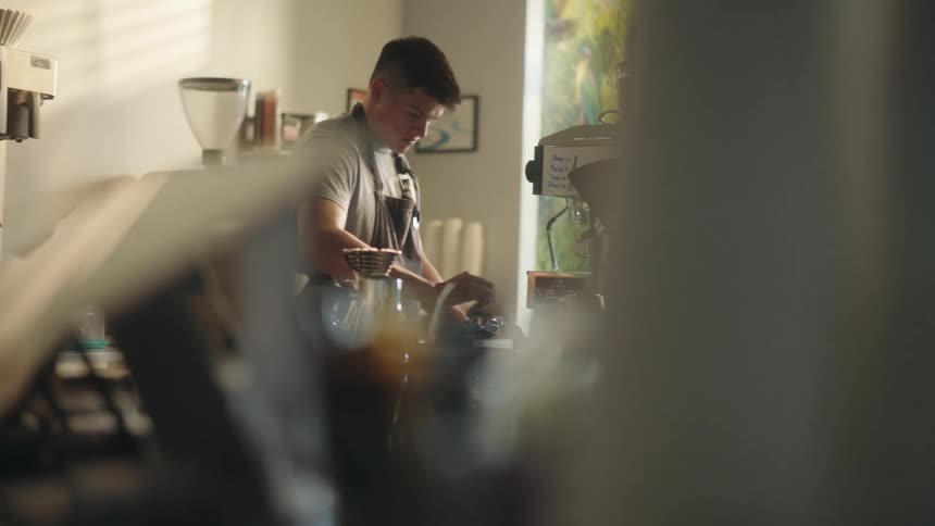 A young male barista grinds coffee beans and scoops the grinds into a metal cup