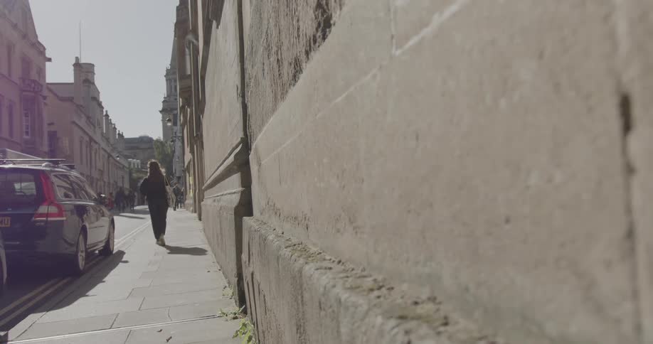 A person walks on a sidewalk beside a stone wall with cars parked on the street