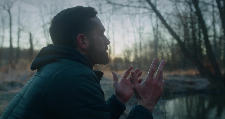 A young man prays by a small body of water in a forest
