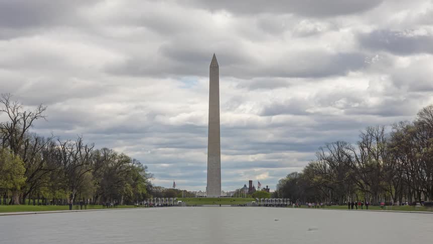 Panorama of a pillar at the washington monument in front of a lake with a pedestrian walking in a cloudy sky