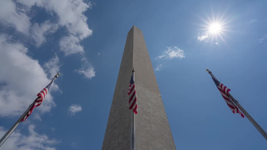 Upward view of a pillar with us flags around and the sky with clouds passing by in time lapse