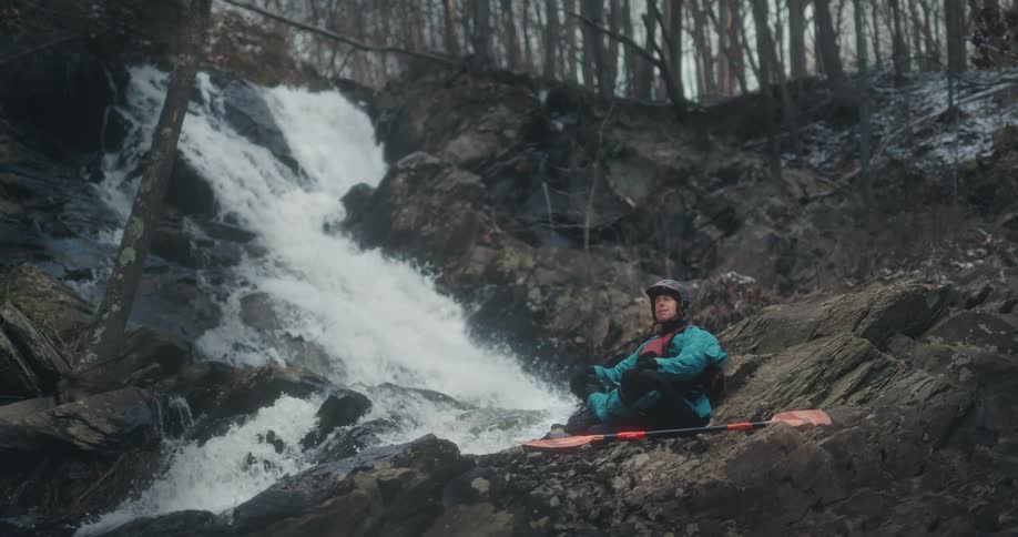Man sitting on a rock next to a waterfall in nature with canoeing equipment and helmets
