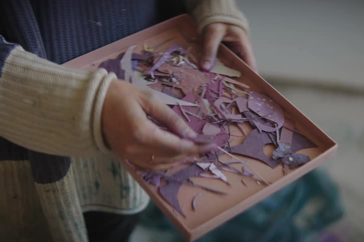 A person is holding a cardboard box filled with rectangular scrap pieces of purple paper