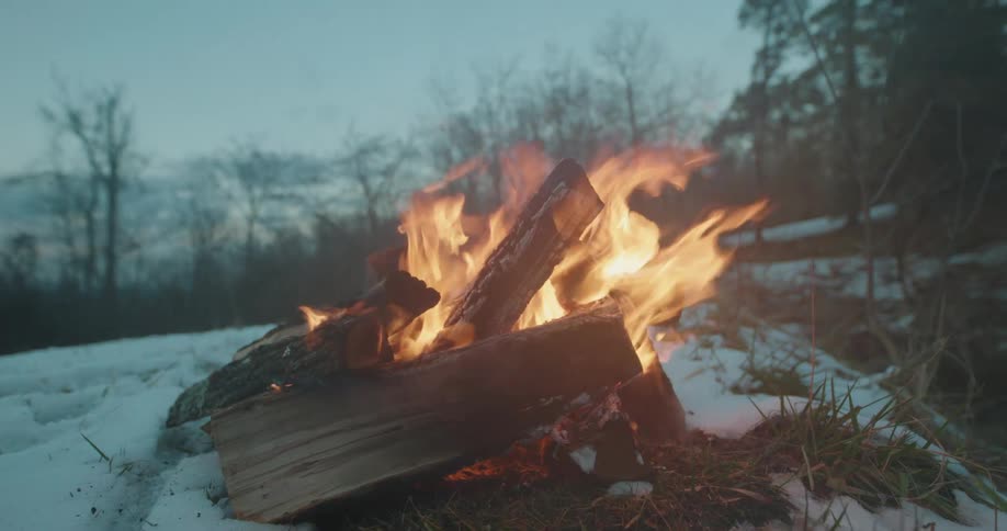 Campfire burns on a snow covered hill with a forest of bare trees in the distance