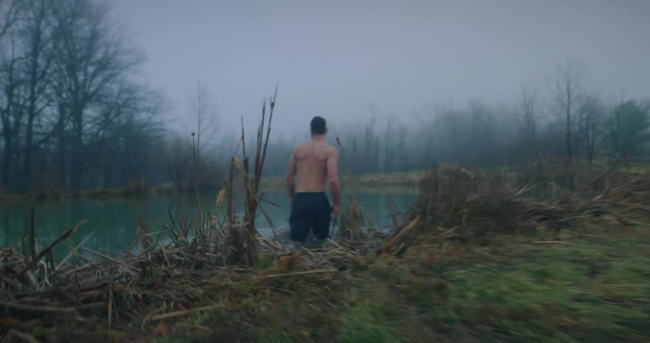 A shirtless man enters a lake surrounded by tall grass in a foggy morning