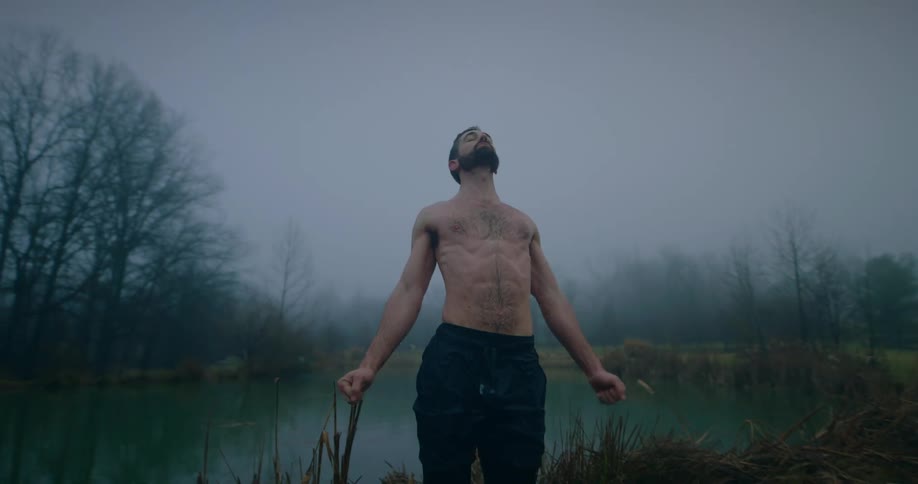 A shirtless man stretching his chest and behind him there is a lake with a little fog behind