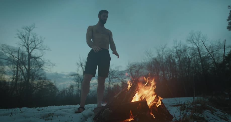 Man without a shirt warming himself standing in front of a fire in a snowy forest on a morning