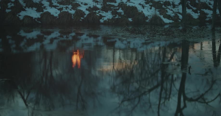 Lake reflecting trees of a forest and a burning bonfire with snow around