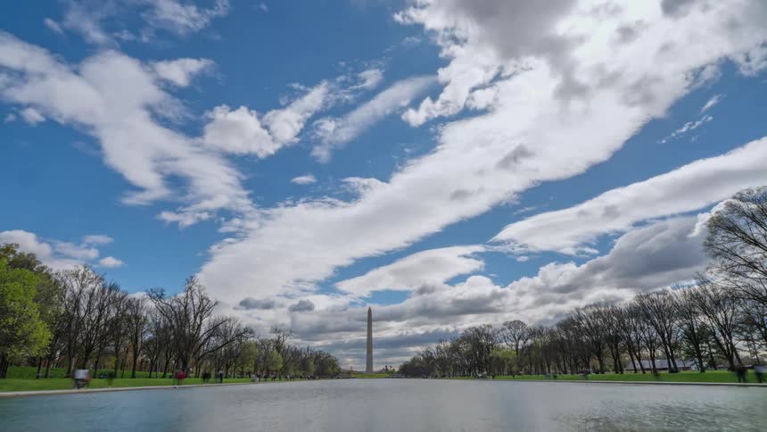 Panorama of a square with pedestrians walking around a lake and in the background a pillar and a day passing by with clouds in timelapse