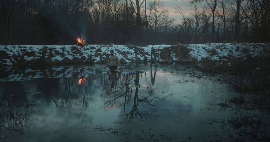 Man without a shirt entering a lake in the forest surrounded by snow