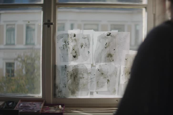 Woman walks through art studio with painted sheets of paper taped onto window