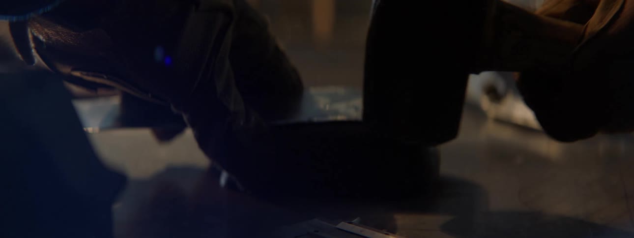 Person wearing gloves hammers thin narrow sheet on workbench in a workshop