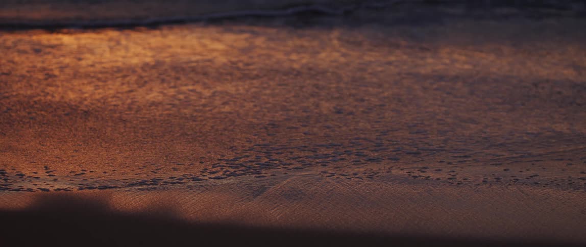 Slow motion footage of ocean waves receding from a beach at sunset