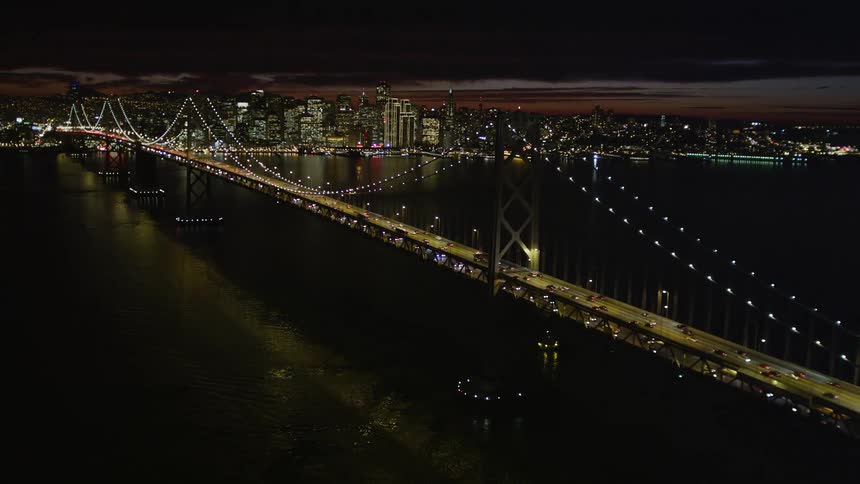 Aerial panorama of a long bridge illuminated at night with vehicle traffic and skyscrapers on the horizon