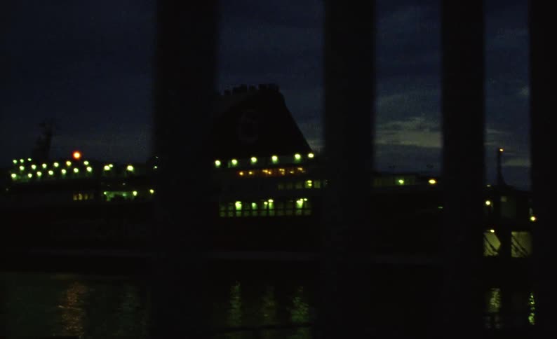A ship parked in front of water that is reflecting the surrounding environment and is dark with some lights on