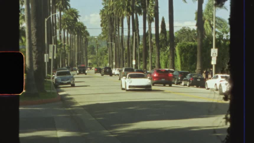 Traffic drives along street lined with palm trees and parked vehicles