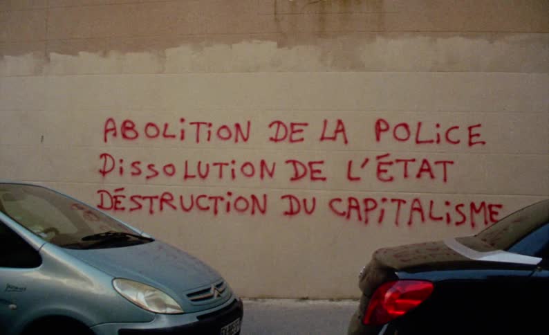 Street in a city with parked cars and a wall with red graffiti with protest writing