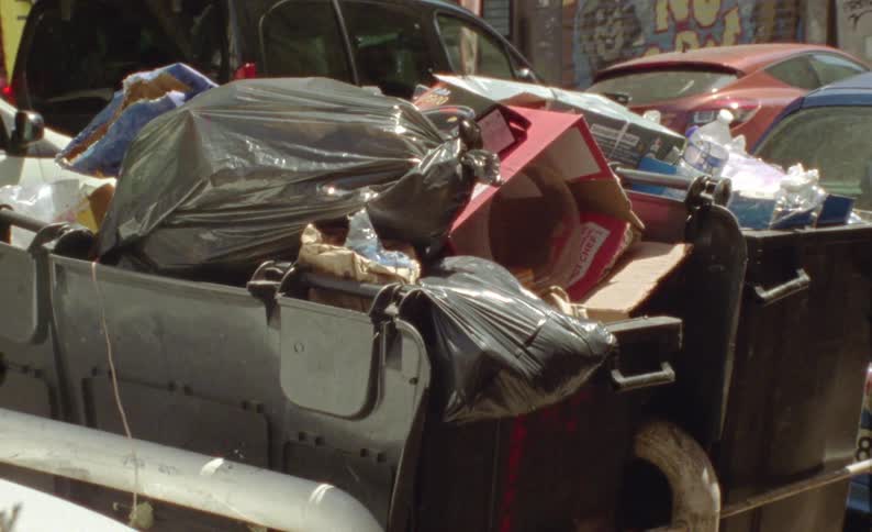 A dumpster that is overflowing with trash and behind it are some parked cars