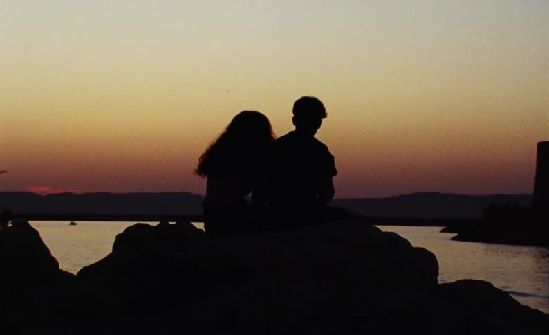 A silhouette of two people sitting on a rock facing a bay