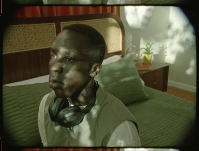 Youth wearing headphones around his neck sits on bed as outdoor plants cast shadows