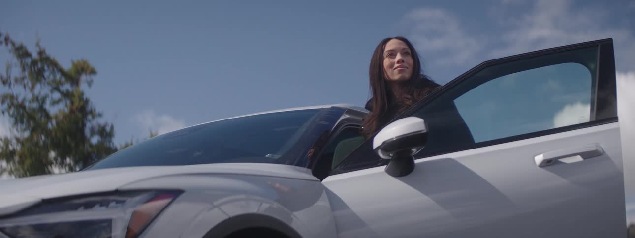 Woman smiles while standing between car and open car door on a cloudy day