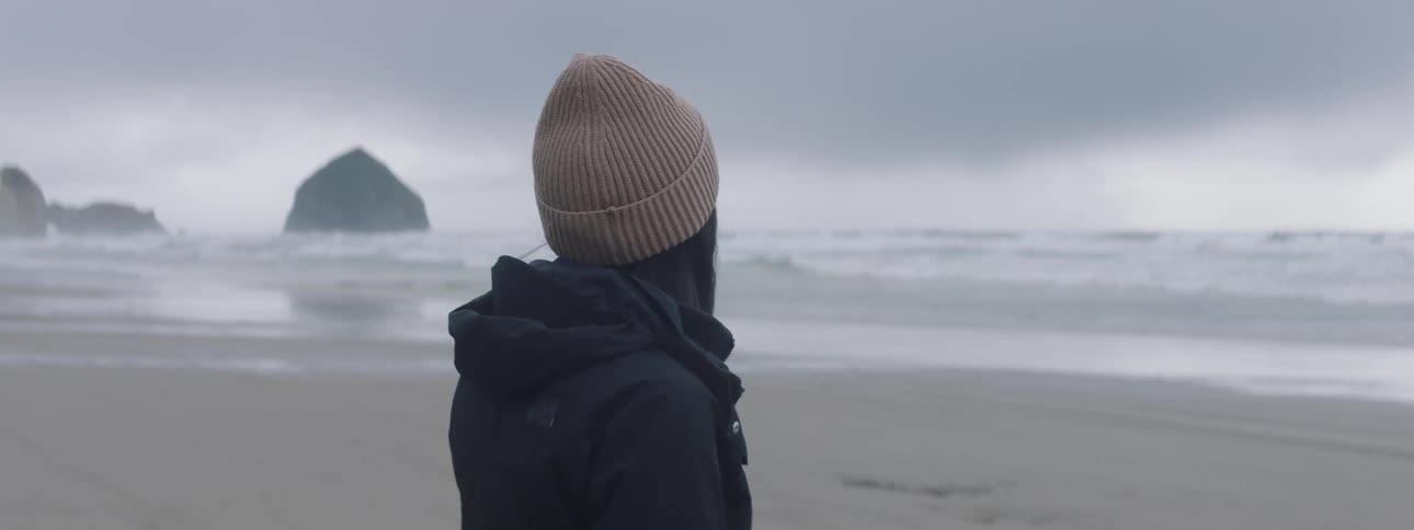 A woman in a striped beanie and black jacket facing the ocean with a rock formation in the distance