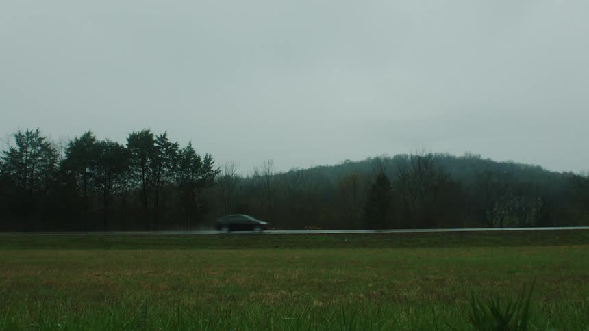 Black car passing through a paved road in the middle of fields and trees around and indian on the side