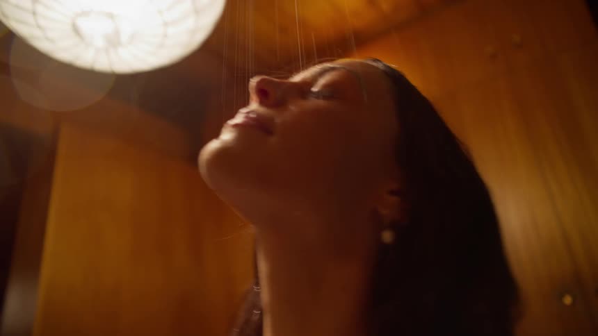 Woman lifts her head as water showers from above in wood paneled room