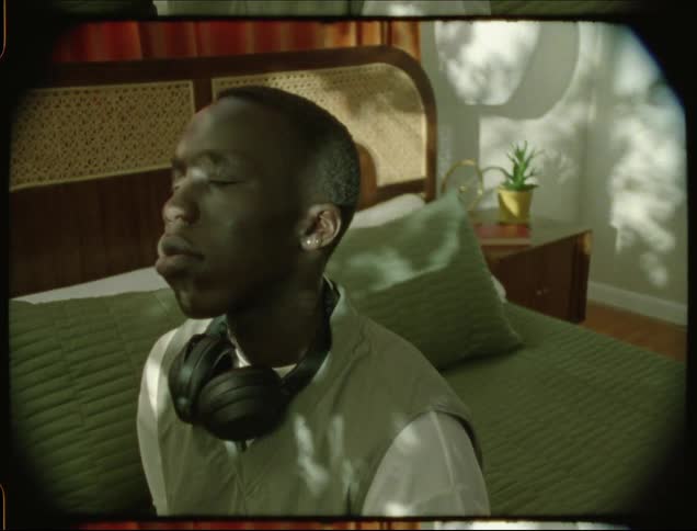 A black man with headphones sitting on a bed with his eyes closed