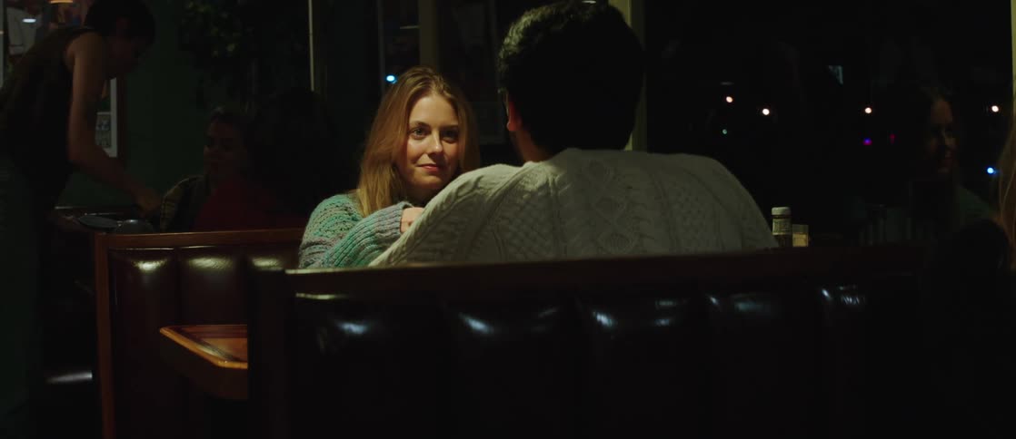 A couple eats a meal together in a busy restaurant the woman smiles then talks to her partner as he sips his drink