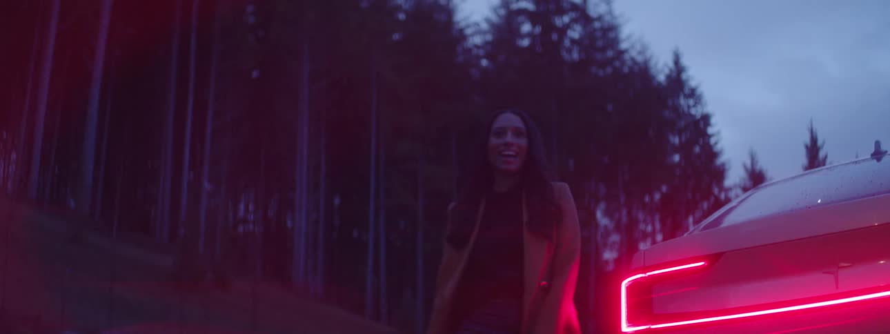A smiling woman walks out and behind a car parked at the side of a forest road