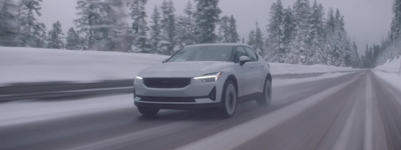 A white car driving on a snowcovered road with several trees on the sides