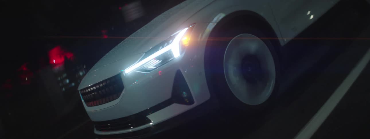 Front and side of a white car with its headlights on driving at night through a city