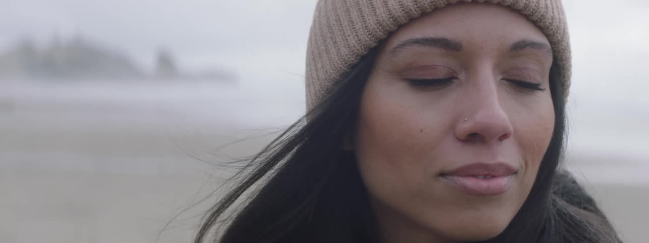 A woman in a striped beanie facing the ocean with a rock formation in the distance