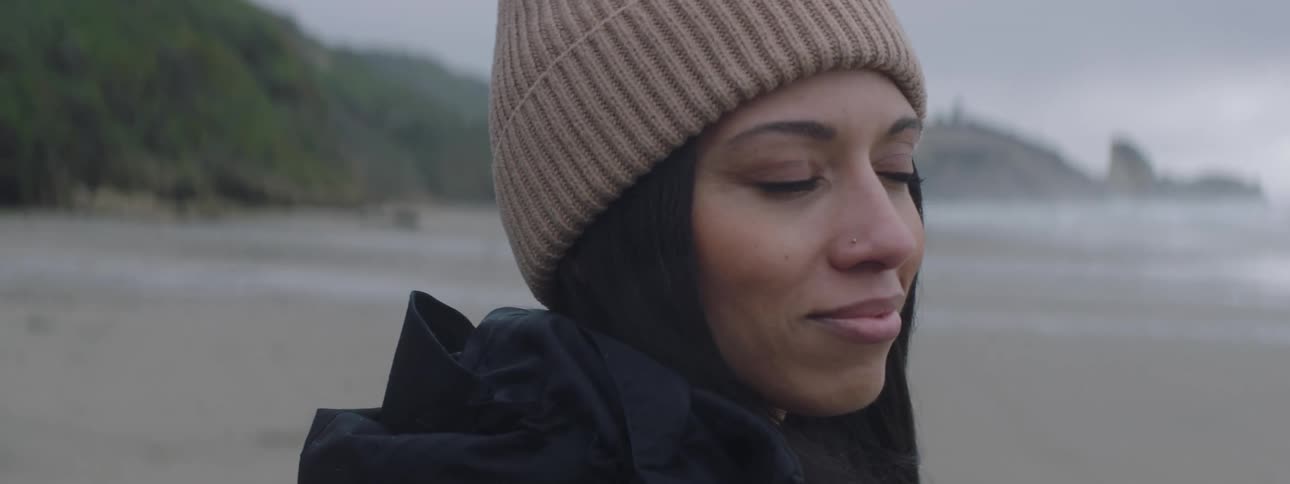 A woman in a striped beanie and black jacket facing the ocean with a rock formation in the distance