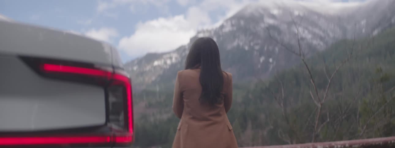 Woman passing by the back of a parked car and overlooking snowy mountains and forests during the day