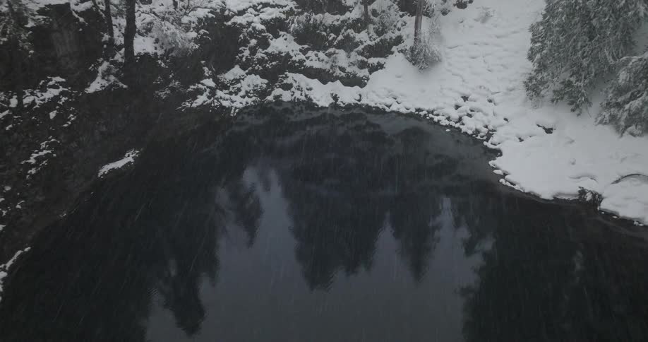 Snow falls onto dark reflective pond surrounded by conifer forest during winter
