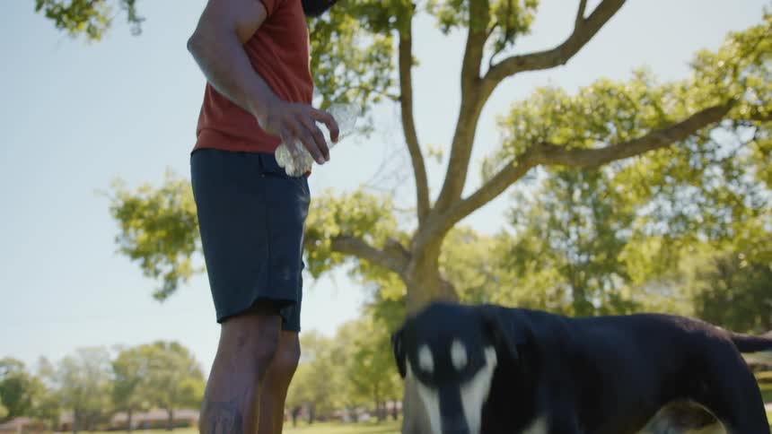 Man holding a bottle of water and offering it to his dog while they are in a park on a clear day