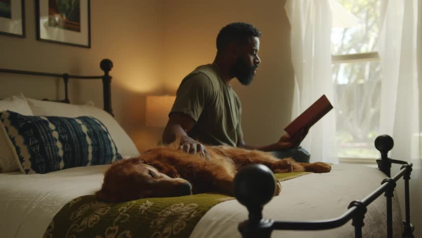 Man reading a book sitting on the bed next to his sleeping dog