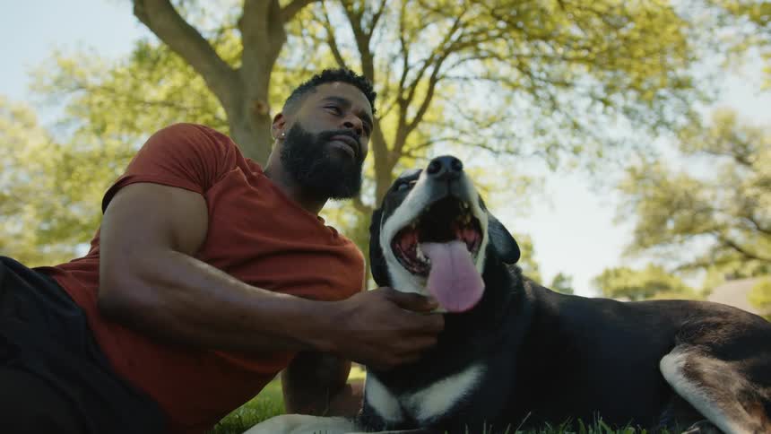 A bearded man is lying on the floor petting his black dogs neck