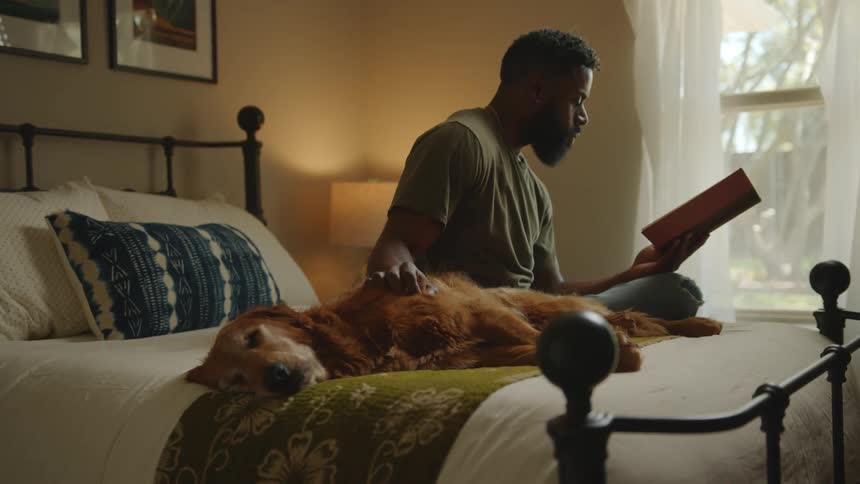 A man reads a book while his dog rests next to him in a bed