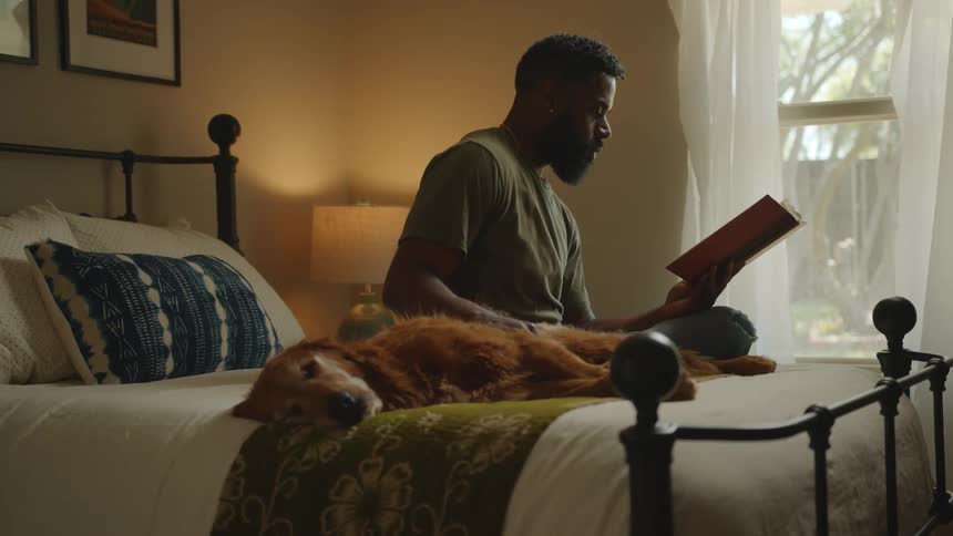 A man reads a book in bed next to a resting dog