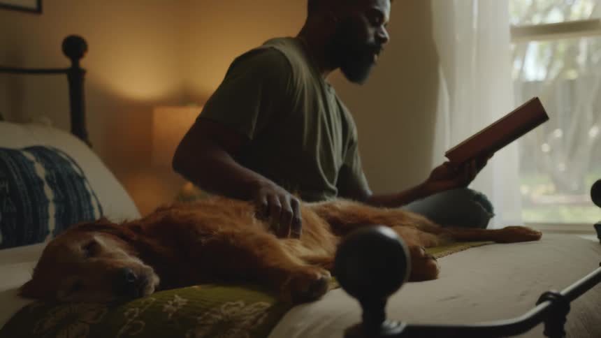 A black man reads in bed while petting his resting dog