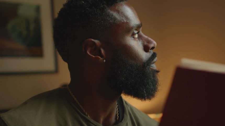 A black man looks away from reading a book in a room
